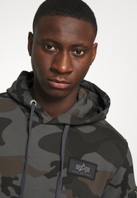 Álcázás mintájú kapucnis pulóver, szürke és barna árnyalatokkal, húzózsinórral ellátott kapucnival és fekete Alpha Industries logós patch-csel. Textúrált anyag.