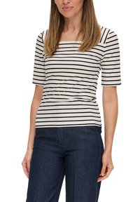 Vrouw in een witte short-sleeve shirt met zwarte horizontale strepen en donkerblauwe jeans, staande met de armen ontspannen langs haar zij.