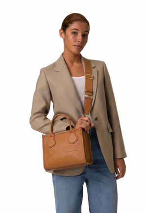 Femme portant un blazer beige et un jean bleu, tenant un sac à main en cuir marron clair de la marque Lancel avec une large bandoulière, sur fond blanc.