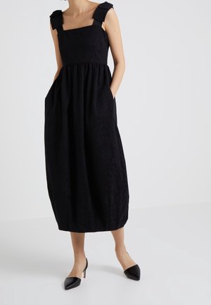 Femme portant une robe midi noire sans manches avec de larges bretelles et des poches, associée à des escarpins noirs à bout pointu.