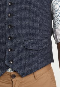 Marineblauwe tweed vest met een gestructureerd patroon, zwarte knopen en een enkele zijzak, gecombineerd met kaki-kleurige broek en een hectisch shirt.