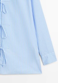 Chemise à manches longues rayée verticalement bleu clair et blanc avec des liens latéraux et des manches retroussées sur fond blanc.