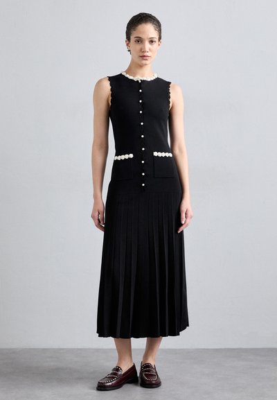 Robe tricotée noire à design sans manches, col échancré, accents de fleurs blanches et poches avant. Jupe plissée s'étend jusqu'au mollet.