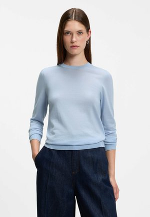 Jeune femme aux cheveux bruns et raides portant un pull bleu clair à manches longues et un jean large bleu foncé, debout les mains dans les poches.