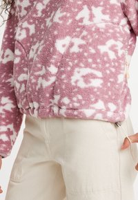 Fleece-Pullover in zartem Rosa mit weißem abstraktem Muster, ausgestattet mit einer Reißverschlusstasche und Raffung am Saum. Kombiniert mit beigen Hosen.