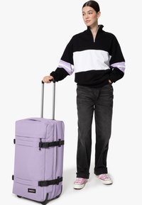 Fialový kufr na kolečkách s dvěma černými popruhy a rukojetí, obdélníkový tvar, vyrobený z odolné textilie, umístěný vedle osoby v neformálním oblečení.