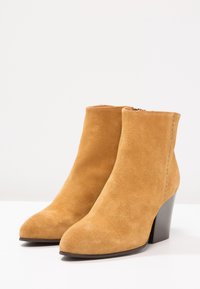 Botines de ante en mostaza con puntera afilada y tacón rectangular. Presentan detalles de costura a lo largo del costado para añadir textura.