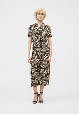 Donna con capelli biondi ricci che indossa un abito midi beige e nero a stampa zebrata con maniche corte e sandali neri con tacco alto.