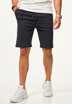 Mann trägt schwarze Shorts mit Kordelzug, weiße Sneaker, weißes T-Shirt, eine Uhr am linken Handgelenk und Armbänder am rechten Handgelenk vor einfarbigem Hintergrund.