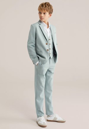 Lyseblåt jakkesæt med en figursyet jakke, treknaps vest og matchende bukser. Hvid skjorte og hvide sneakers giver kontrast.