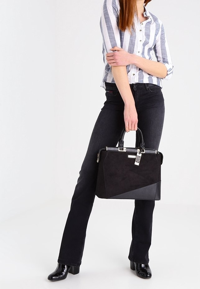 Femme portant une chemise à rayures boutonnée et un pantalon noir, tenant un sac à main en cuir et daim noir par ses poignées.