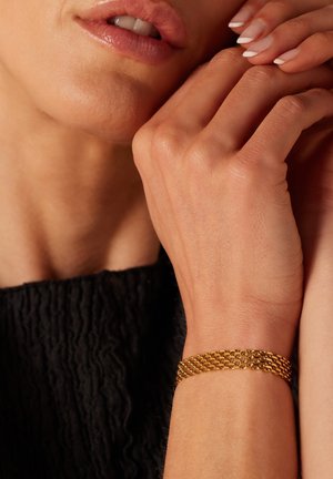 Gouden geweven armband om een pols, met een gestructureerd ontwerp. De hand is gedeeltelijk bedekt, met korte, verzorgd nagels.