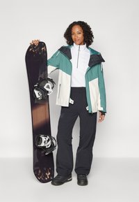 Snowboard har en blank svart ovansida, med bindningar fästa. Individuen bär en jacka i teal, vit och svart färg, med matchande byxor och stövlar.