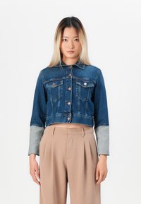 Woman with straight long hair wearing a cropped denim jacket with gray cuffs and high-waisted beige trousers, standing against a white background.