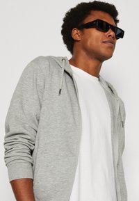 Gray zip-up hoodie with drawstring, white t-shirt beneath, and black rectangular sunglasses. Fabric appears soft; casual fit.