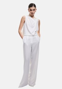 Top bianco senza maniche con un design drappeggiato e un accent in oro sulla spalla, abbinato a pantaloni a gamba larga con vita alta nello stesso tessuto.
