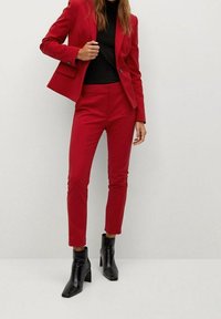 Nainen, joka on pukeutunut punaisiin räätälöityihin pukuhaalareihin, joissa on kapeat housut, mustaan poolopaitaan ja mustiin korkeat nilkkasaappaisiin, seisoo vaaleaa taustaa vasten.