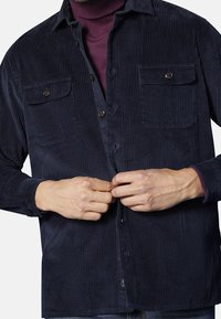 Camisa de pana en color marino con costillas verticales, dos bolsillos en el pecho y cierres de botón. Mangas largas con detalle en los puños y un brillo sutil.