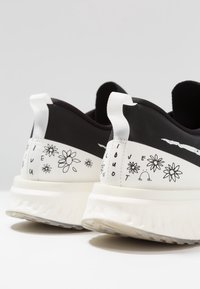 Černobílé tenisky s květinovými vzory a textem na patě. Mají polstrovanou bílou podrážku a kontrastní černý svršek.