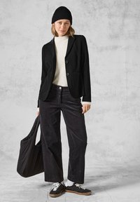 Černý blazer s dvěma knoflíky, nošený přes bílé žebrované svetr. Spárovaný s tmavými kordurovými kalhotami a černou čepicí. Drží černou tašku.