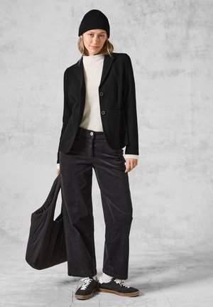 Černý blazer s dvěma knoflíky, nošený přes bílé žebrované svetr. Spárovaný s tmavými kordurovými kalhotami a černou čepicí. Drží černou tašku.