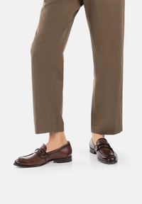 Brown leather loafers with a smooth finish, featuring a decorative strap and buckle, paired with light brown, slightly tapered trousers.