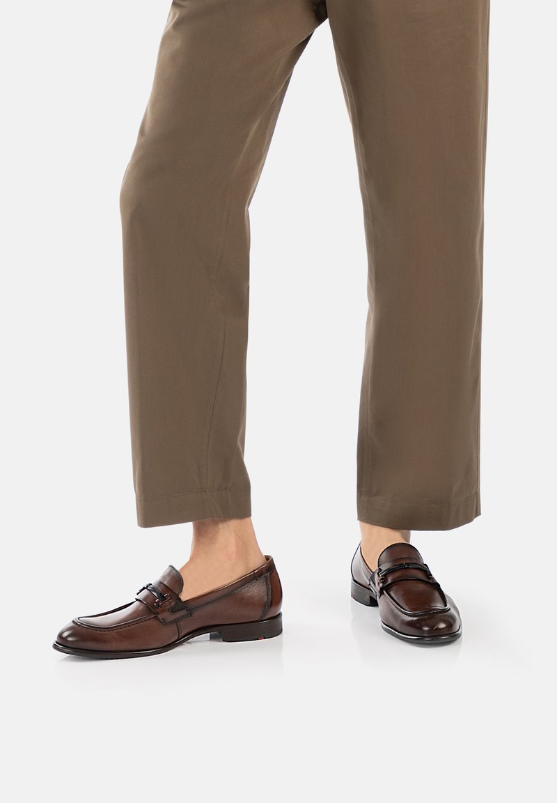 Brown leather loafers with a smooth finish, featuring a decorative strap and buckle, paired with light brown, slightly tapered trousers.