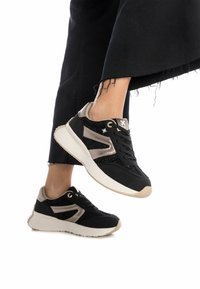 Zapatillas negras con tela texturizada, acentos dorados y una suela de color claro. Presentan un diseño de cordones y franjas laterales en contraste.