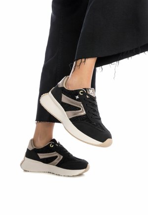 Zapatillas negras con tela texturizada, acentos dorados y una suela de color claro. Presentan un diseño de cordones y franjas laterales en contraste.