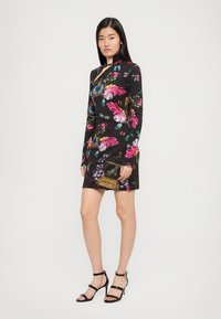 Zwart langemouwen jurk met kleurrijke bloemen- en vlinderpatronen, met een aansluitend ontwerp en een lengte tot midden boven de dij. Geaccentueerd met een gouden handtas.