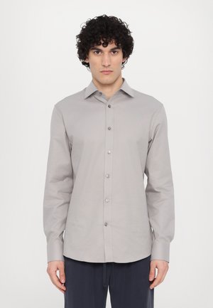 Chemise grise à manches longues avec un col à boutons, présentant un plastron avant avec des boutons métalliques et un design à coupe décontractée.
