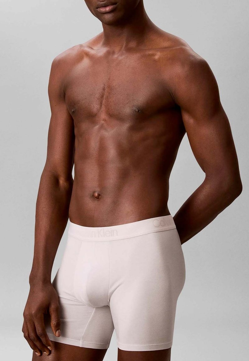 Fit donkerhuid mannelijk torso, gekleed in lichtbeige Calvin Klein boxershorts, staand met één arm achter zijn rug tegen een effen achtergrond.