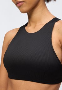 Záber zblízka na ženu, ktorá nosí čierny rebrovaný krátky top bez rukávov s vysokým golierom na jednofarebnom pozadí.