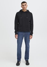 Črna jopa s kapuco in vrvicami, nošena čez jeans hlače. Ima sproščen kroj in rebraste manšete, kombinirano s črnimi supergami.