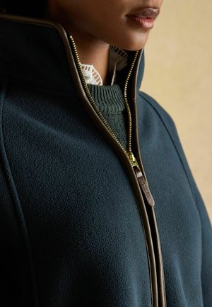 Fleece jacket in dark teal with a high collar and brown zipper. Textured fabric with a zippered front, layered over a green knit sweater.