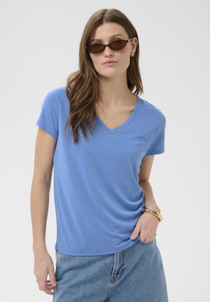 Jonge vrouw met bruine zonnebril, een blauwe T-shirt met V-hals, spijkerbroek, gouden oorringen en een gouden armband, poserend met haar hand in haar zak.