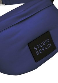 Plavi najlon fanny pak s crnim logom koji glasi "STUDIO BERLIN." Ima glatku teksturu i podesivu traku za sigurno prianjanje.