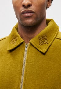 Volnena jakna v gorčično rumeni barvi z zadrgo, okrašena z vezenimi cvetličnimi vzorci na ovratniku in teksturirano mehko površino.