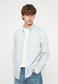 Νέος με καστανά σγουρά μαλλιά, φοράει ένα ανοιχτό μπλε πουκάμισο πάνω από λευκό T-shirt και μπλε τζιν, κοιτάζοντας προς τα πλάγια.