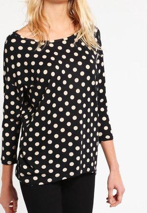 Maglia nera a maniche lunghe con grandi pois color crema. Presenta un ampio scollo e una texture morbida. Indossata con pantaloni neri.