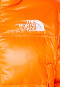 Jasnopomarańczowa kurtka puchowa z białym haftowanym logo "The North Face" na klatce piersiowej oraz widocznymi detalami przeszycia.