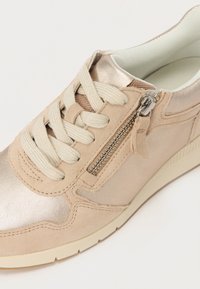 Béžové tenisky s kombinací semiše a metalických akcentů, obsahují tkaničky, zipy a texturované detaily, navrženy s polstrovanou podrážkou.