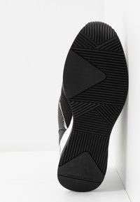 Semelle de basket noire avec motifs géométriques texturés et logo "Michael Kors", présentée sur une surface plane blanche dans un coin.