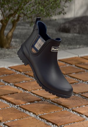 Botte noire en caoutchouc jusqu'à la cheville avec des accents à carreaux, semelle texturée et logo de marque. La botte présente une finition lisse et des panneaux latéraux élastiques.