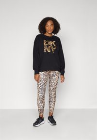 Zwarte sweatshirt met een luipaardprint logo, gecombineerd met leggings met luipaardprint. Model draagt zwarte sneakers. Eenvoudige achtergrond.