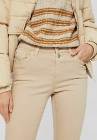 Béžové slim fit džíny s dvěma předními kapsami a kovovými knoflíky, v kombinaci s pruhovaným béžovým a hnědým pleteným svetrem a lehkou péřovou bundou.