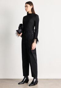 Sieviete valkā melnu ribotu garroku blūzi ar spalvu aprocēm, melnas augsta jostasvieta bikses, melnas potīšu zābaciņus un tur melnu rokassomiņu.