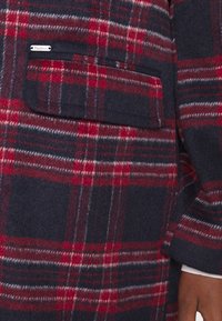 Manga de abrigo de lana de cuadros en rojo, azul marino y blanco con bolsillo de solapa y pequeña etiqueta metálica "Papa Jeans", combinado con puño visible de camisa blanca.