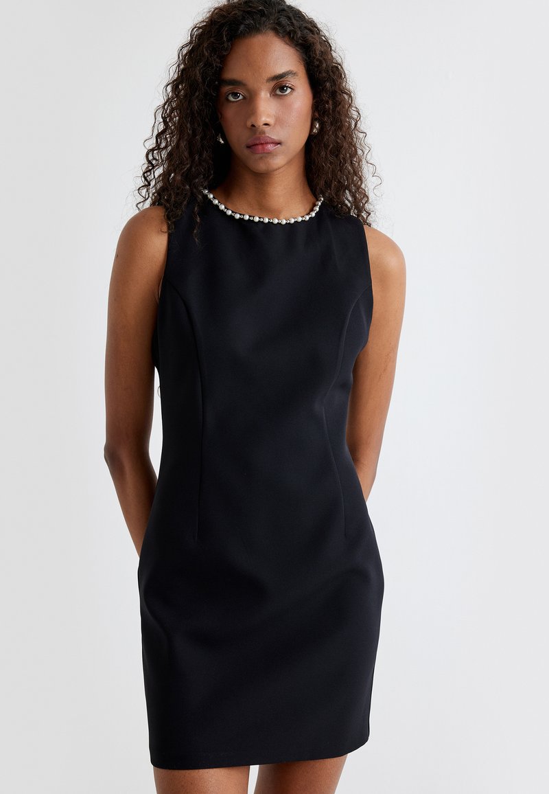 Vestido negro sin mangas con un diseño ajustado, que presenta un escote adornado con perlas y una textura de tela suave, de longitud hasta la rodilla con costuras mínimas.
