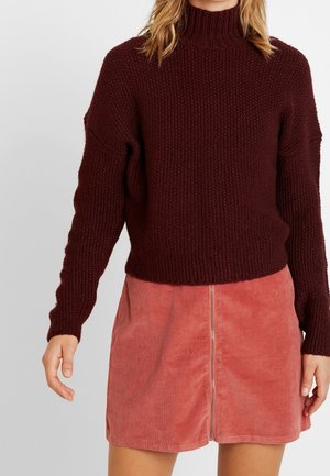 Femme portant un pull en laine rouge foncé et une jupe en velours côtelé rose avec une fermeture éclair à l'avant, debout devant un fond clair uni.